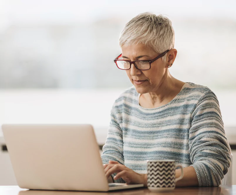 A woman sits at a table typing on a laptop, coffee mug by her side. The Silver & Archibald disability lawyers have secured more in Social Security Disability benefits than any Georgia-based law firm.